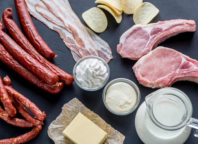 Tabletop with foods including, sausage, bacon, pork, cream, butter and potato chips.