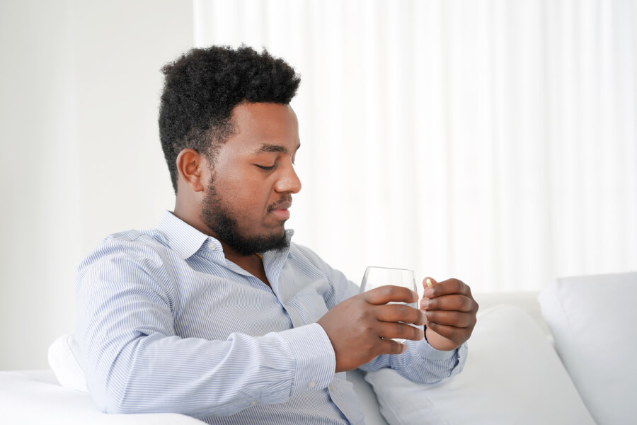 Man sitting on a couch taking a pill with a glass of water.