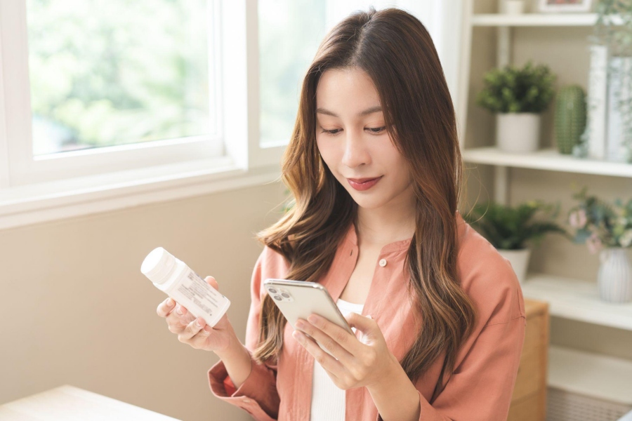 Young woman reading supplement label on phone in cozy room