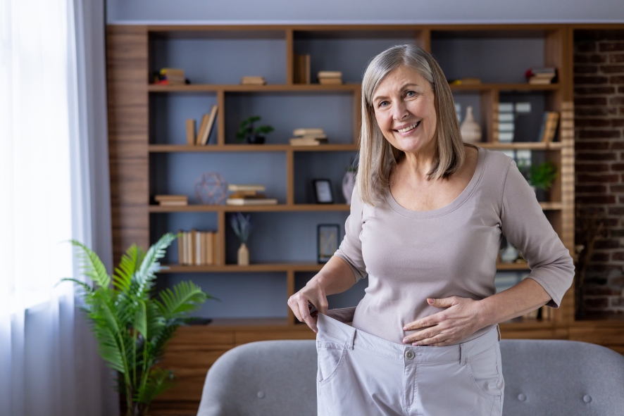 Smiling mature woman showing weight loss success standing indoors holding large pants in relaxed setting